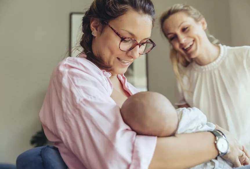 Mother breasfeeding a human baby child while another person is happily looking at them.