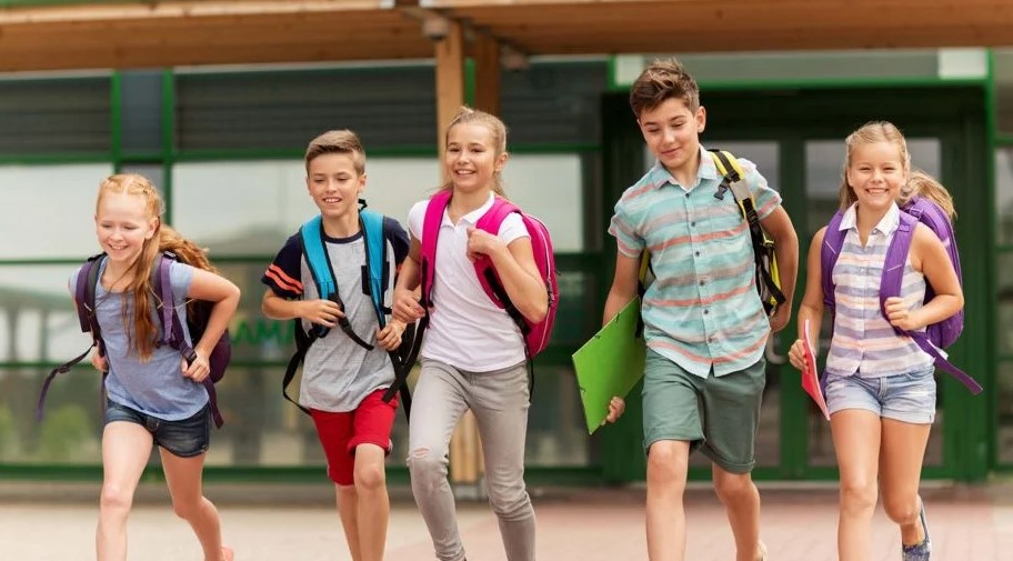 Image of 5 children wearing backpacks walking in front of a school