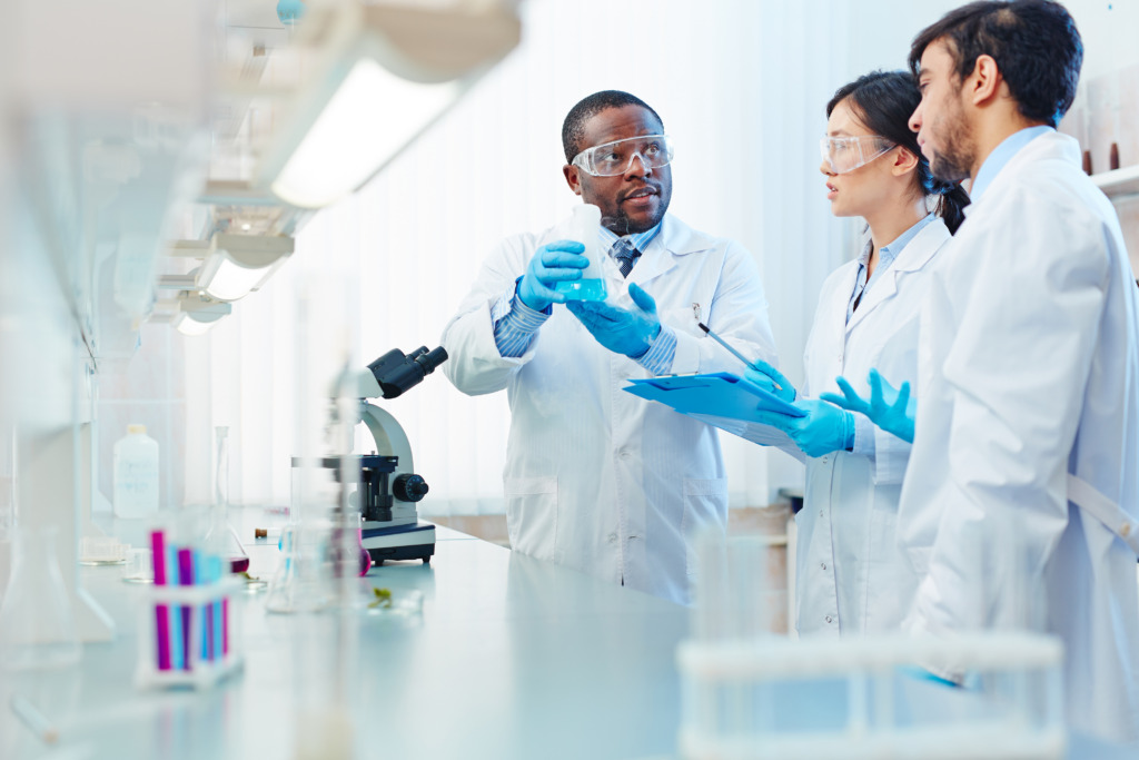 Male African-American laboratory scientist holding flask with blue liquid discussing chemical reaction with male Latin-American and female Asian colleagues.